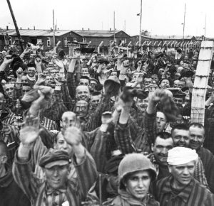 Black and white Dachau liberation photograph. Men wearing striped prison uniforms cheer in front of and behind barb-wire fence. One American soldier stands in front.
