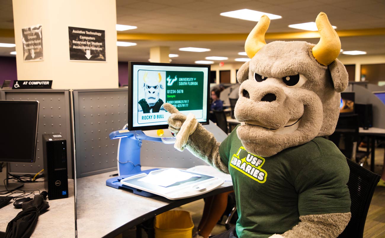 USF mascot Rocky sitting at an image enlarger in the Tampa campus library. Rocky is facing the camera, pointing at the screen in front of him, which is showing an enlarged photo of his USF ID.