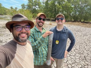 three men smile at the camera while standing in a forested area with cracked earth under their feet and green tress in the background