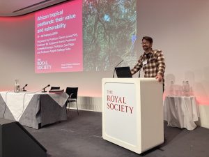 Chris Kiahtipes stands at a podium with the word Royal Society printed on it, preparing for a presentation which is projected behind him.