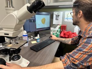 Chris Kiahtipes looks at biological material on a computer screen. there is a large microscope next to the computer.