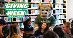 Rocky the Bull stands with a group of students at the USF Tampa campus Library, with a USF Giving Week logo at the top left corner.