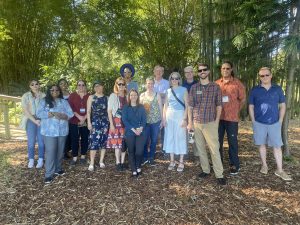 A group of 14 people smiling at the camera. The people are outdoors in a woodsy setting.