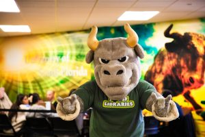 USF mascot Rocky D. Bull does horns with his hands in front of a bull mural inside a library.
