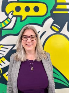 A woman with long hair and glasses is wearing a purple top with a grey blazer and stands, smiling, in front of what looks like a mural. it is dark green, bright green, and yellow.