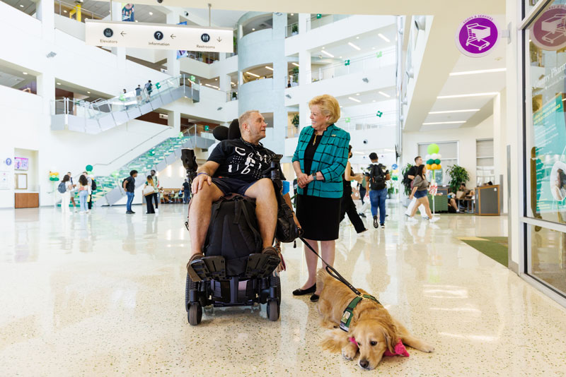 USF president Rhea Law chats with a man in a motorized chair, along with his service dog, in the Marshall Student Center